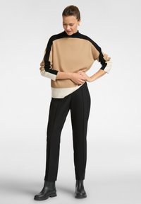 Beigefarbener Strickpullover mit schwarz-weißen Akzenten, hohem Kragen und oversize Passform. Maßgeschneiderte schwarze Hosen und schwarze Ankle-Boots runden den Look ab.