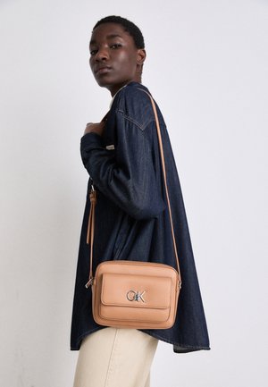 Person trägt eine dunkle Jeansjacke und helle Hose, trägt eine beige Umhängetasche mit "CK" Logo auf der Vorderseite der Tasche.