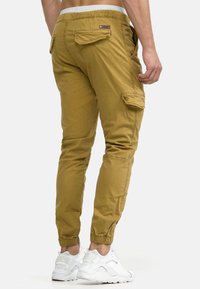 Khakifarbene Cargo-Hosen mit schmaler Passform, elastischen Bündchen und mehreren Taschen. Hergestellt aus leichtem Material. Kombiniert mit weißen Sportschuhen.