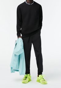 Sweat-shirt noir avec col à logo, pantalon noir, tenant une veste bleu clair. Baskets vert néon avec accents en maille et semelles texturées.