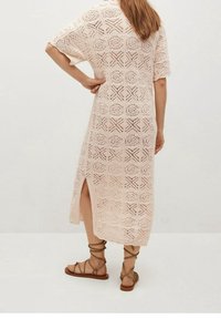 Robe en crochet longue de couleur rose clair avec des motifs carrés élaborés et des manches courtes. Une fente sur le côté dévoile une partie de la jambe. Portée avec des sandales plates.