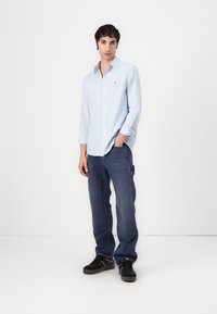 Cămașă cu butoni, de culoare albastră deschisă, cu dungi albe și portocalii, asociată cu pantaloni cargo de culoare albastră închisă și adidași negri. Design simplu și casual.