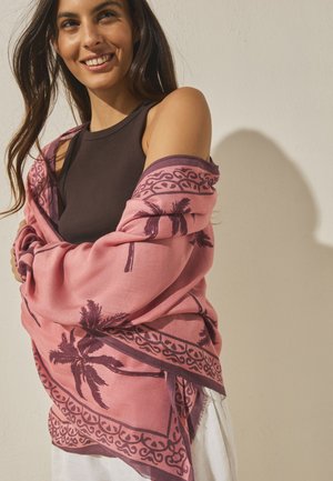 Usmievavá žena s dlhými tmavými vlasmi v čiernom tielku bez rukávov a s ružovým šálom s tmavými vzormi palmových listov.