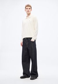 Gaiši bēša garām piedurknēm polo krekls ar sarkanu logotipu, kopā ar platu tumši zilu džinsu biksēm. Melni ādas apavi noslēdz tērpu.