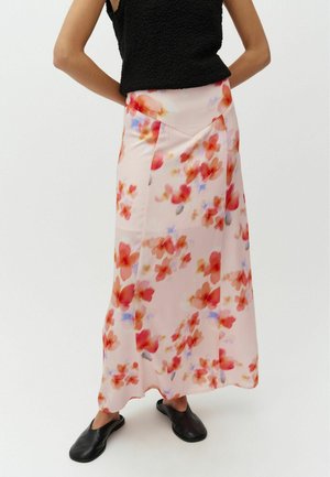 Femeie purtând o fustă lungă, roz deschis, cu imprimeu floral roșu și mov, top negru fără mâneci, cu textură, și pantofi slip-on negri.