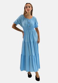 Jasnoniebieska sukienka maxi z krótkimi bufkowymi rękawami, marszczonym stanem, detalami na guziki oraz falbanami na spódnicy; zestawiona z czarnymi balerinami.