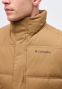 Άνδρας που φορά γαλακτώδες μπουφάν Columbia με ψηλό γιακά, ορατό αυτί με μικρό ασημένιο κρίκο, ουδέτερο φόντο.