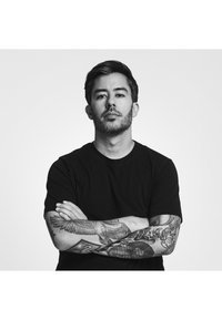 Svart t-shirt på en person med tatuerade armar. Tatueringarna har intrikata mönster, inklusive fåglar och abstrakta former. Neutral bakgrund.