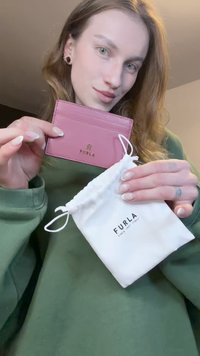 Roza usnjena denarnica za kartice z navpičnimi režami, vtisnjenim logotipom; skupaj z belo vrečko z vrvico, na kateri je črno napisano blagovno znamko.