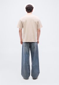 Άνδρας που στέκεται με την πλάτη στραμμένη, φοράει μπεζ πουκάμισο με κοντά μανίκια και φαρδιά μπλε τζιν, μπροστά σε απλό ανοιχτό φόντο.