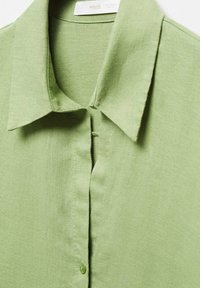 Camisa verde clara con cuello, cierre frontal de botones y tejido texturizado. Presenta un material suave y transpirable con un diseño informal.