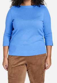 Pull en tricot léger bleu, à manches trois-quarts et à encolure ronde. Associé à un pantalon marron en suède. Texture unie sur l'ensemble.