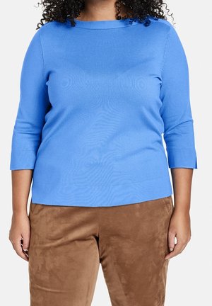Blauer, leichter Strickpullover mit Dreiviertelärmeln und rundem Halsausschnitt. Trägt dazu eine braune, suedenähnliche Hose. Ganze Zeit schlichtes Muster.