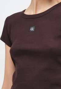 Camiseta de algodón burdeos con textura acanalada y cuello redondo. Cuenta con una etiqueta cuadrada negra con logo blanco en el lado izquierdo. Mangas cortas.