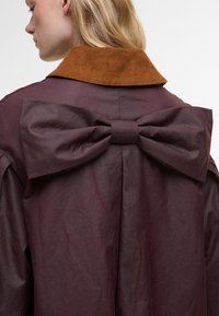 Μπουργουντί παλτό με καφέ χοντρό κολάρο, με λεπτομέρεια μεγάλης κορδέλας πίσω. Λείο ύφασμα, υπερμεγέθης σιλουέτα.