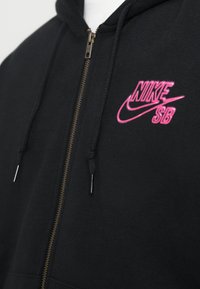 Čierna mikina na zips s ružovým logom Nike SB na ľavej hrudi a čiernymi šnúrkami so kovovými koncami.