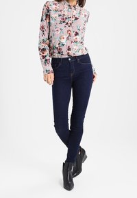 Camicia a bottone floreale con motivi rosa, rossi e verdi; abbinata a jeans skinny blu scuro; indossata con stivali alla caviglia neri.