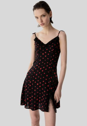 Giovane donna che indossa un vestito nero senza maniche con spalline sottili, motivo floreale rosso e rifiniture in pizzo su uno sfondo grigio chiaro.