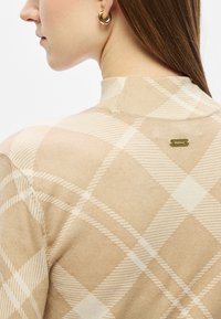 Pulover bej cu un model ușor în carouri, cu guler înalt și etichetă cu logo auriu pe spate. Textură netedă, croială mulată.