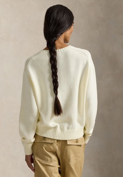 Πουλόβερ σε κρεμ απόχρωση, πλεκτό με ριμπ τελείωμα στη μέση και στα μανίκια, σε άνετη εφαρμογή και με διακριτή υφή. Φορεμένο με μπεζ παντελόνια, εμφανίζει πλεξούδα στα μαλλιά.