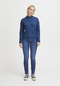 Modrý, texturovaný sveter s vysokým golierom a dlhými rukávmi, skombinovaný s priliehavými modrými džínsami a čiernymi teniskami. Jednoduchý, neformálny outfit.