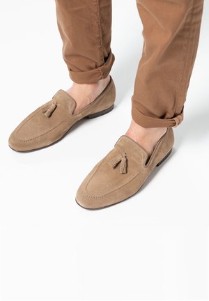 Mocassins en daim beige clair avec un léger éclat. Dotés d'un gland décoratif et de détails de couture. Bout arrondi et semelle à profil bas.