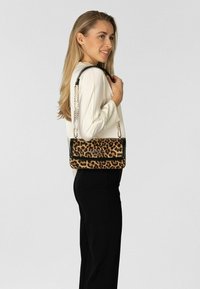 Kvinde i hvid bluse og sorte bukser bærer leopardmønstret håndtaske med guldkæde over skulderen, smilende og kigger til siden.