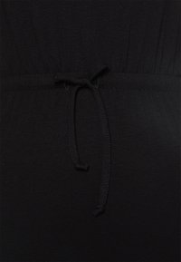 Černá látka zblízka na šatech s elastickým pasem a stahovací šňůrkou, která vytváří skládací efekt. Měkká textura s minimálním leskem.