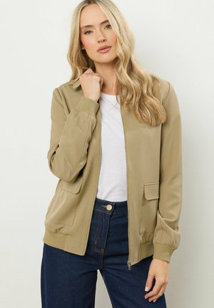 Beige Bomberjacke mit Reißverschluss, gerippten Manschetten und Taschen. Kombiniert mit einem weißen Hemd und hoch taillierten, dunklen Jeans. Angenehme Textur.
