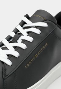 Baskets noires Tommy Hilfiger avec des lacets blancs et un logo doré sur le côté et la languette, montrant un gros plan détaillé de la partie supérieure.
