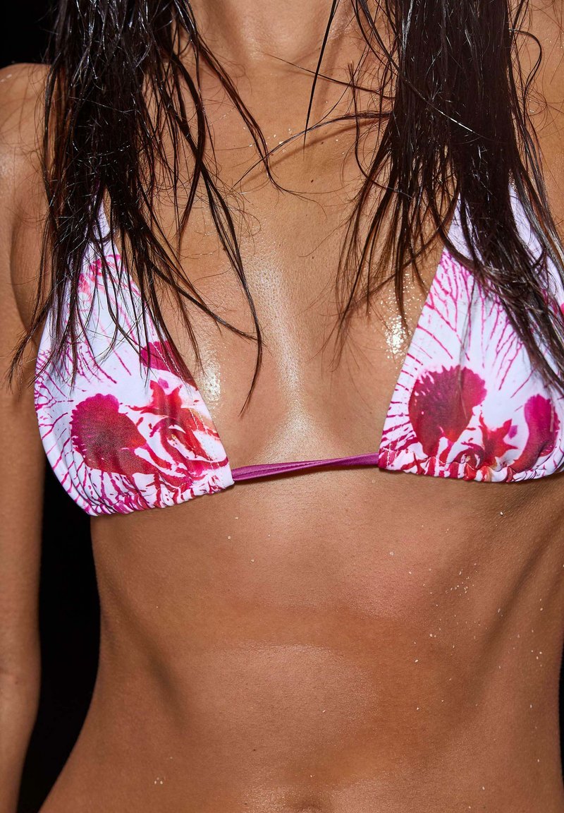 Bikini top s trojúhelníkovými košíčky z bílé látky s červenými květinovými vzory. Tenké fialové ramínka. Pleť vypadá leskle a mokře.