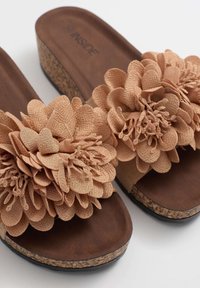 Chanclas beige con suelas de cuña de corcho y grandes acentos de flores de tela en las correas. Textura suave con una plantilla lisa de color marrón oscuro.