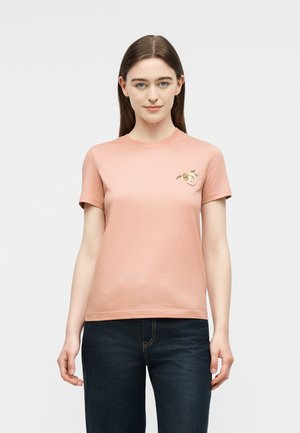 Fiatal nő egyenes barna hajjal, barackszínű rövid ujjú pólót visel, amelyen virágos hímzés található a mellrészen, valamint sötétkék farmert.