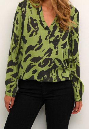 Kvinde iført en grøn bluse med sort abstrakt dyreprint og lange ærmer, kombineret med sorte tætsiddende bukser.