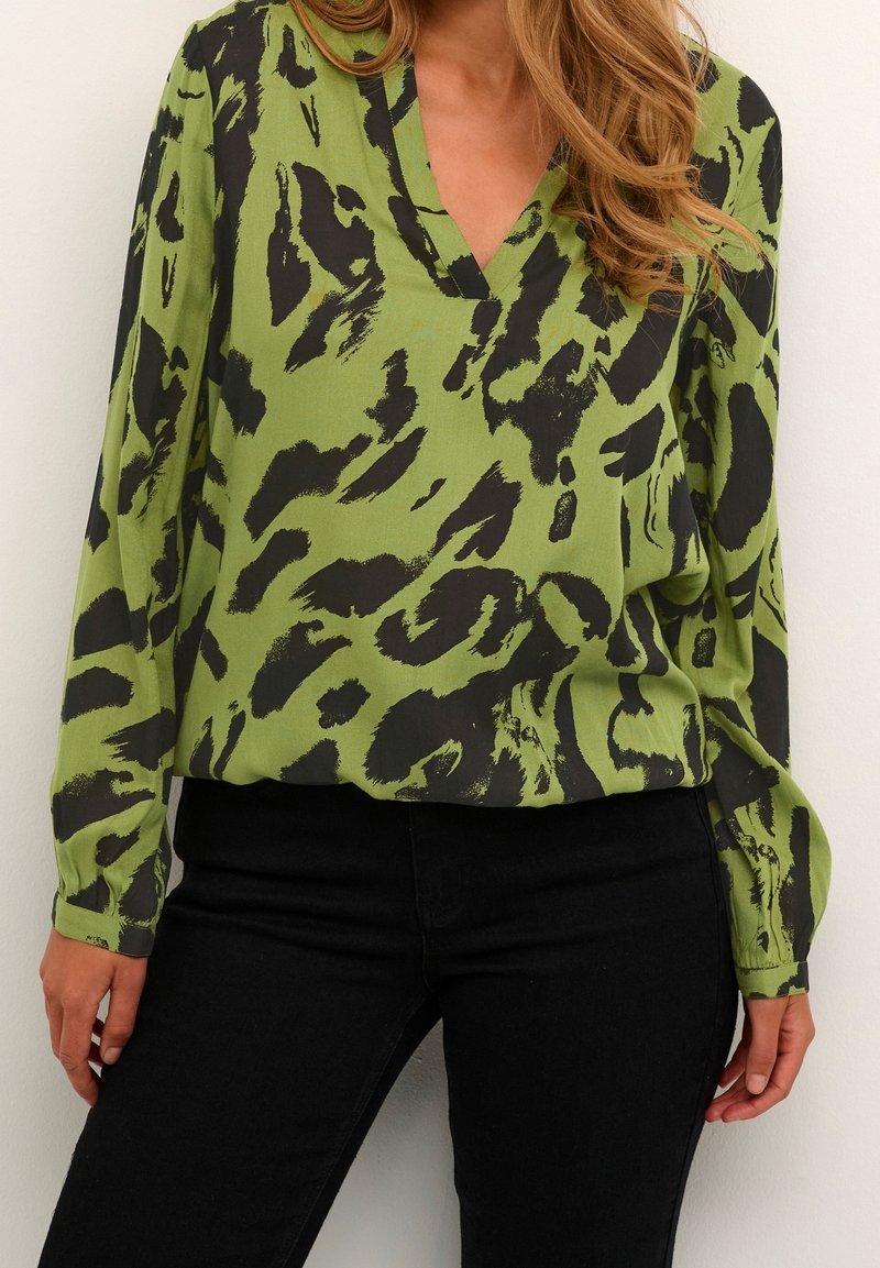 Femme portant un chemisier vert à imprimé animal abstrait noir et manches longues, associé à un pantalon noir ajusté.