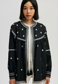 Black embroidered jacket with floral designs, contrasting white stitching, round neckline, and tassel accents at the front. Long sleeves.