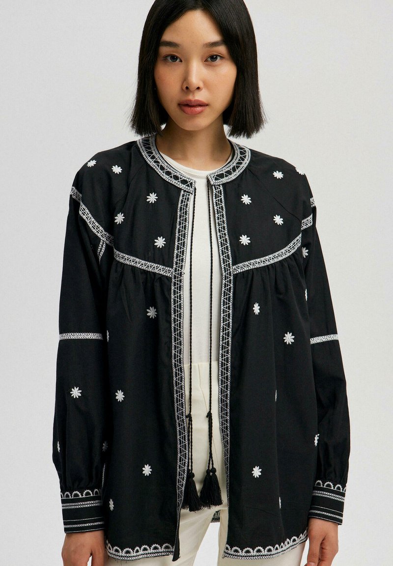 Black embroidered jacket with floral designs, contrasting white stitching, round neckline, and tassel accents at the front. Long sleeves.