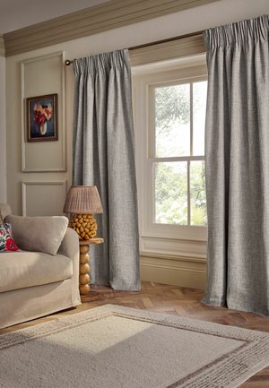 Beige sofa with a floral pillow, a wooden side table with a carved lamp, gray curtains framing a large window, and a textured light rug on wood floor.