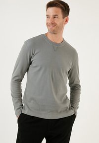 Hosszú ujjú, bordázott felső tompa szürke színben, kerek nyakkivágással és háromszög alakú varrással a gallérnál, sötét nadrággal párosítva.