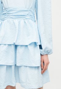 Robe bleu clair texturée avec des volants superposés et de longues manches bouffantes, présentée de la taille aux genoux avec une main visible sur le côté.