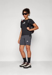 Μαύρο T-shirt με γραφικά, γκρι σορτς, μαύρες κάλτσες και ορειβατικά μποτάκια. Μαύρο σακίδιο με κόκκινες λεπτομέρειες. Γυαλιά ηλίου συμπληρώνουν την εμφάνιση.