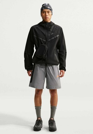 Mann trägt schwarze Reißverschlussjacke, graue Shorts, graue Socken, schwarze Sneakers, Umhängetasche und camouflagefarbene Kopfbedeckung und steht vor weißem Hintergrund.