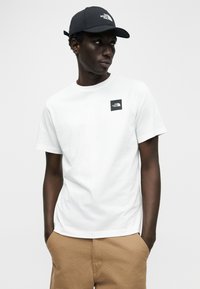 Λευκό βαμβακερό T-shirt με κοντά μανίκια, με ένα μικρό μαύρο ετικετάκι λογοτύπου. Φορώντας το με καφέ σορτς και μαύρο καπέλο.