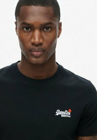 Camiseta de algodón negra con un logo blanco de "SuperDry" y acento rojo. Diseño de cuello redondo. Textura suave y forma ajustada.