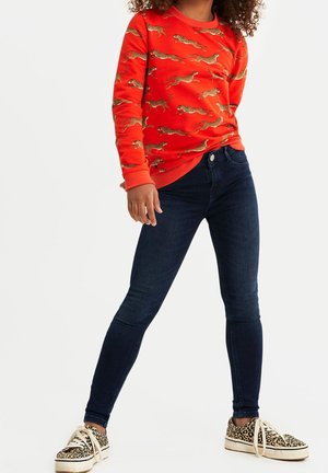 Roter Pullover mit Leopardenmuster, dunkelblaue Skinny-Jeans und Sneakers mit Leopardenmuster. Hat lange Ärmel und einen runden Halsausschnitt.