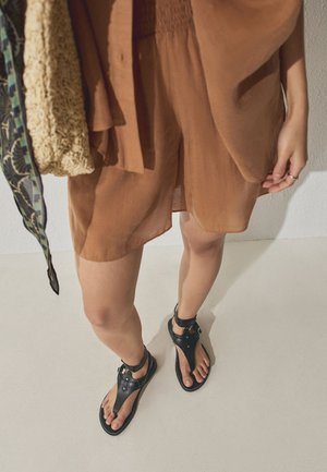 Persona con pantalones cortos marrones y sandalias de cuero negras, sosteniendo una bolsa beige texturizada con una correa con estampado.