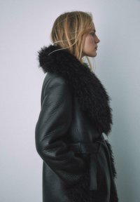 Czarny skórzany płaszcz z dużym, miękkim czarnym futrzanym kołnierzem, długimi rękawami i paskiem wiązanym w talii, o gładkiej fakturze.