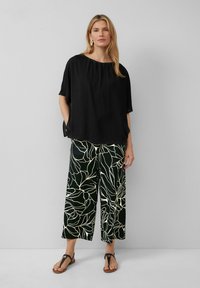 Zwarte vloeiende top met korte mouwen, gecombineerd met brede zwarte broeken met een abstract wit bloemenpatroon. Model draagt een paar bruine sandalen.