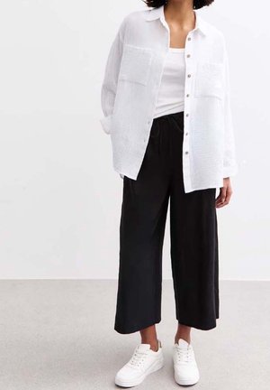 Person wearing a white button-up shirt, white tank top, loose black cropped pants, and white sneakers, standing on a gray floor.