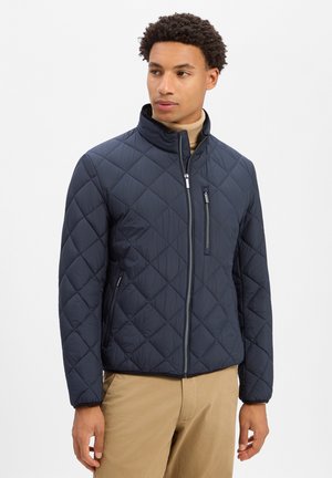 Man draagt een marineblauwe doorgestikte jas met ritssluitingen op de zakken en beige broek, staand tegen een witte, effen achtergrond.